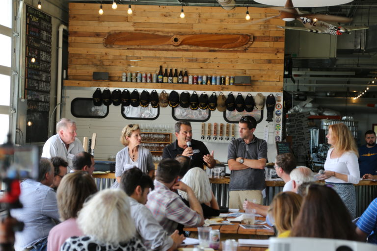 Four speakers and moderator speak to a brewery full of seated individuals