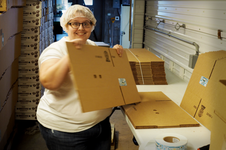 A smiling white woman is holding a cardboard box and smiling