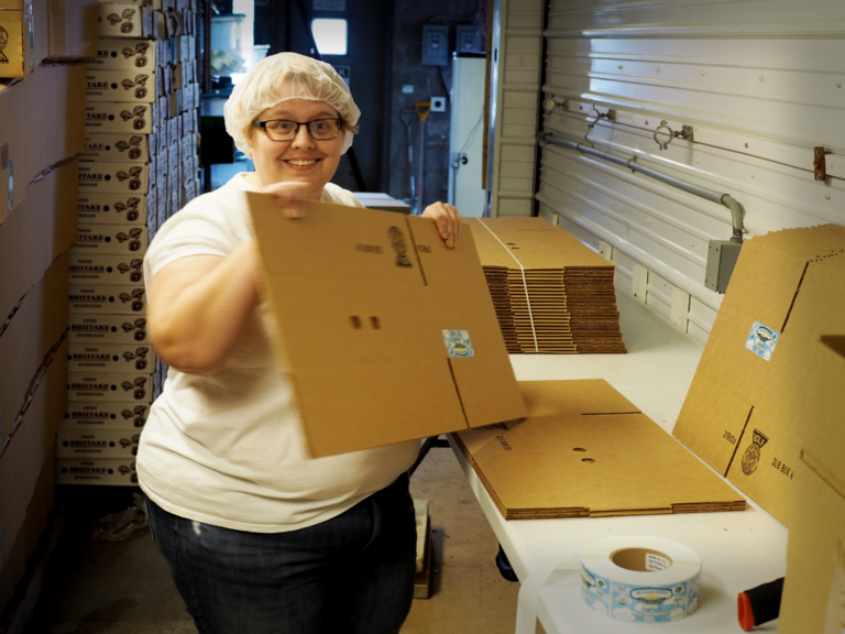 A smiling white woman is holding a cardboard box and smiling