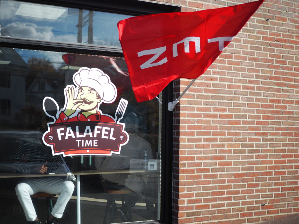 A sign saying Falafel Time with the image of a chef, doing a "chef's kiss" is on the window of a brick building