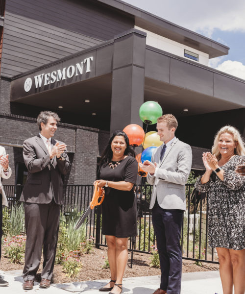 A diverse group of individuals is smiling and clapping as they cut a ribbon in front of a building with the words Wesmont on the front