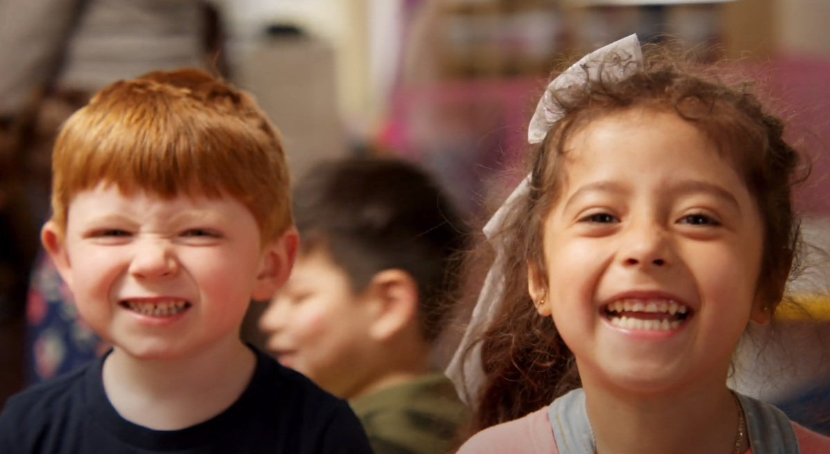 A young white boy with red hair and a Mexican girl, both preschoolers, smile broadly