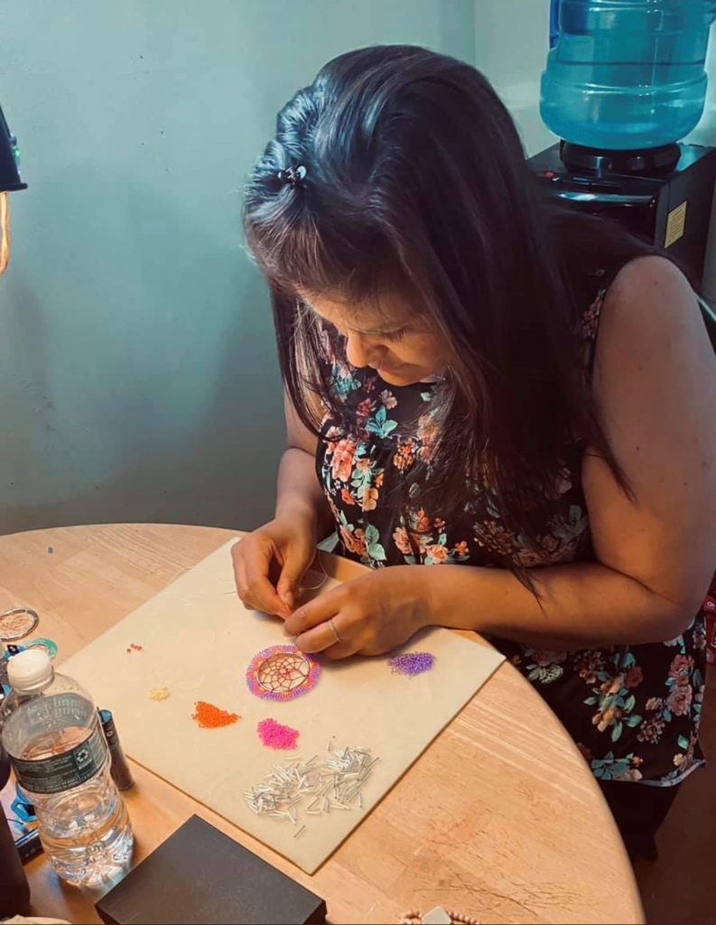 A Passamaquoddy Native American woman is bent over a table making a beaded dreamcatcher