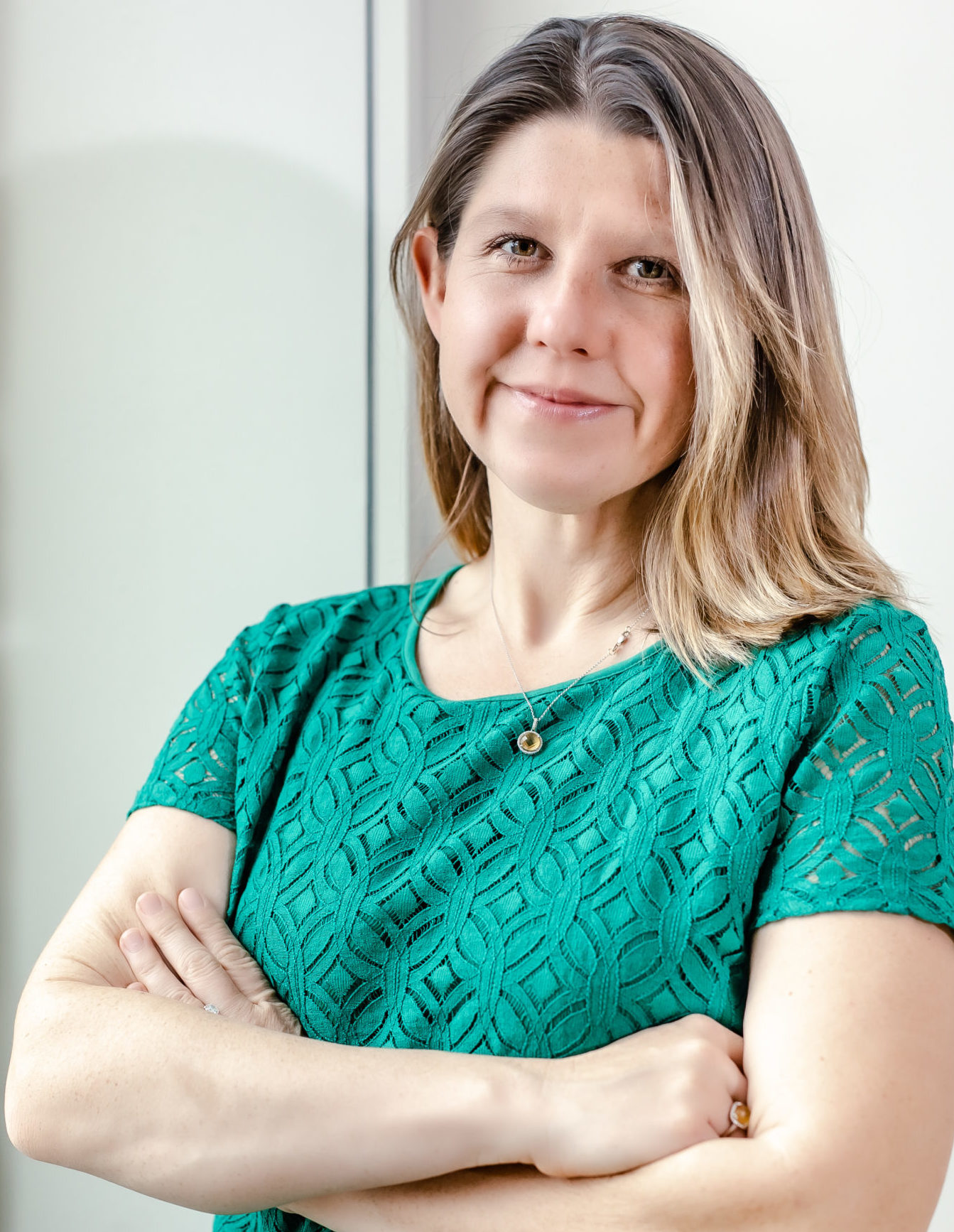 A blonde woman in a teal shirt stands with her arms crossed in front of a white wall