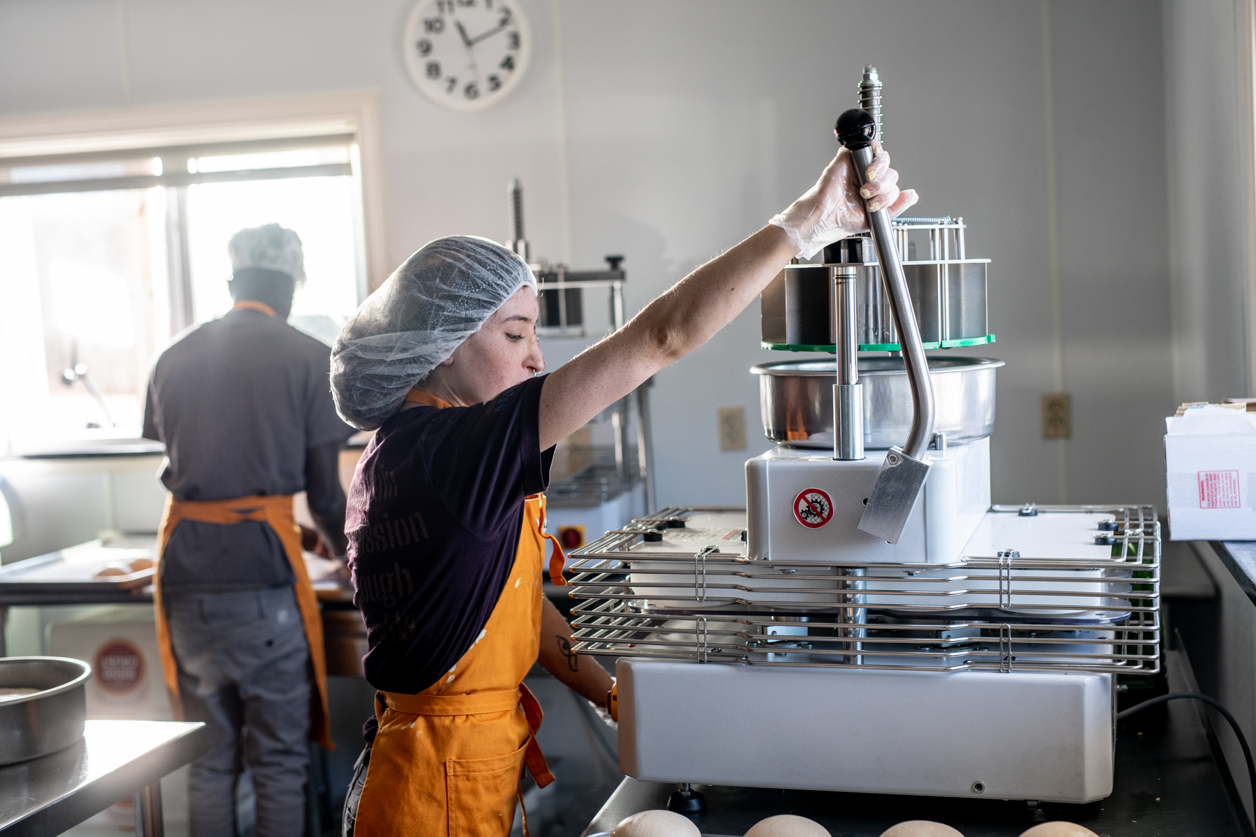 A young white woman in a hairnet packages balls of pizza dough with a machine, while a second employee, a young Black man, works in the background.